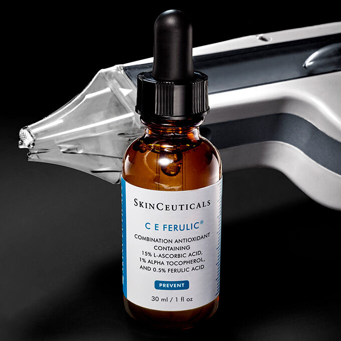 Eine bernsteinfarbene Tropfflasche mit dem SkinCeuticals C E Ferulic Serum ist im Vordergrund positioniert, wahrend ein nicht-ablativer Laser im unscharfen Hintergrund sichtbar ist.