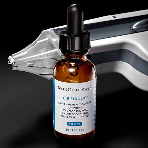 Eine bernsteinfarbene Tropfflasche mit dem SkinCeuticals C E Ferulic Serum ist im Vordergrund positioniert, wahrend ein nicht-ablativer Laser im unscharfen Hintergrund sichtbar ist.