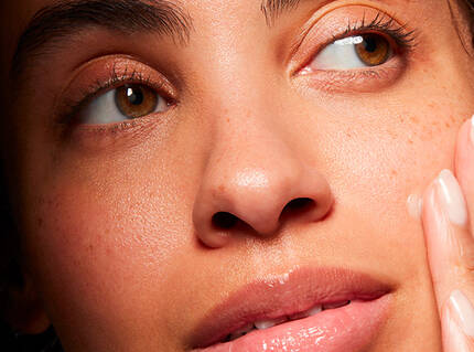 Close-up einer Frau mit braunen Augen und leicht freckeliger Haut, die sanft ihr Gesicht beruhrt.