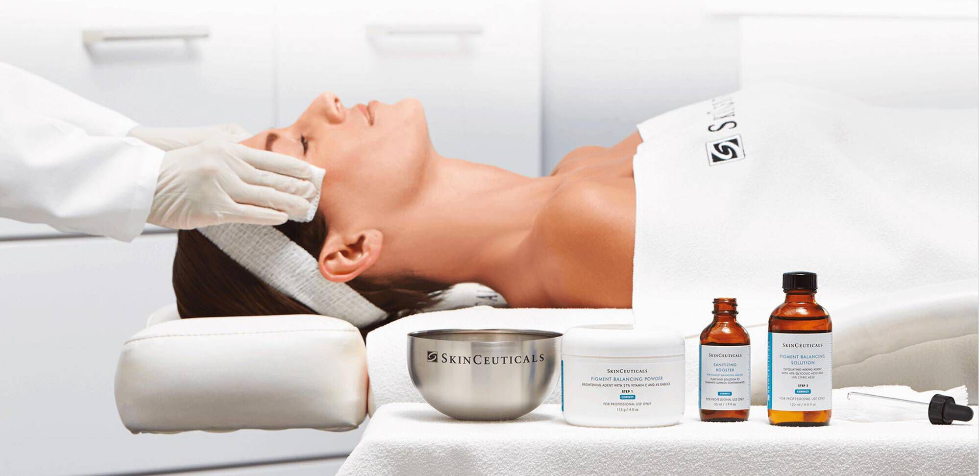 Eine Frau liegt entspannt auf einer Liege mit einer Gesichtsbehandlung. Neben ihr stehen Produkte von SkinCeuticals, darunter Cremes und Seren. Eine Person mit Handschuhen fuhrt die Behandlung durch.
