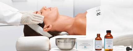 Eine Frau liegt entspannt auf einer Liege mit einer Gesichtsbehandlung. Neben ihr stehen Produkte von SkinCeuticals, darunter Cremes und Seren. Eine Person mit Handschuhen fuhrt die Behandlung durch.