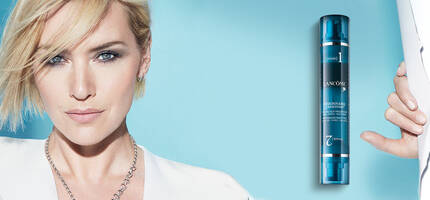 A woman with short blonde hair and a confident expression poses against a blue background, holding a skincare product in her right hand. The product is a sleek bottle labeled with "7".