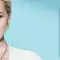 A blonde woman with short hair poses next to a blue skincare product, showcasing a fresh and modern style against a light blue background.