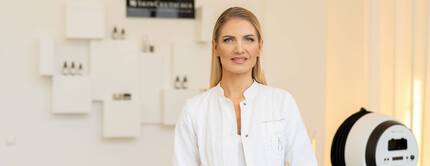 Eine lachelnde Frau in einem weißen Kittel steht in einem modernen Raum mit Hautpflegeprodukten im Hintergrund.