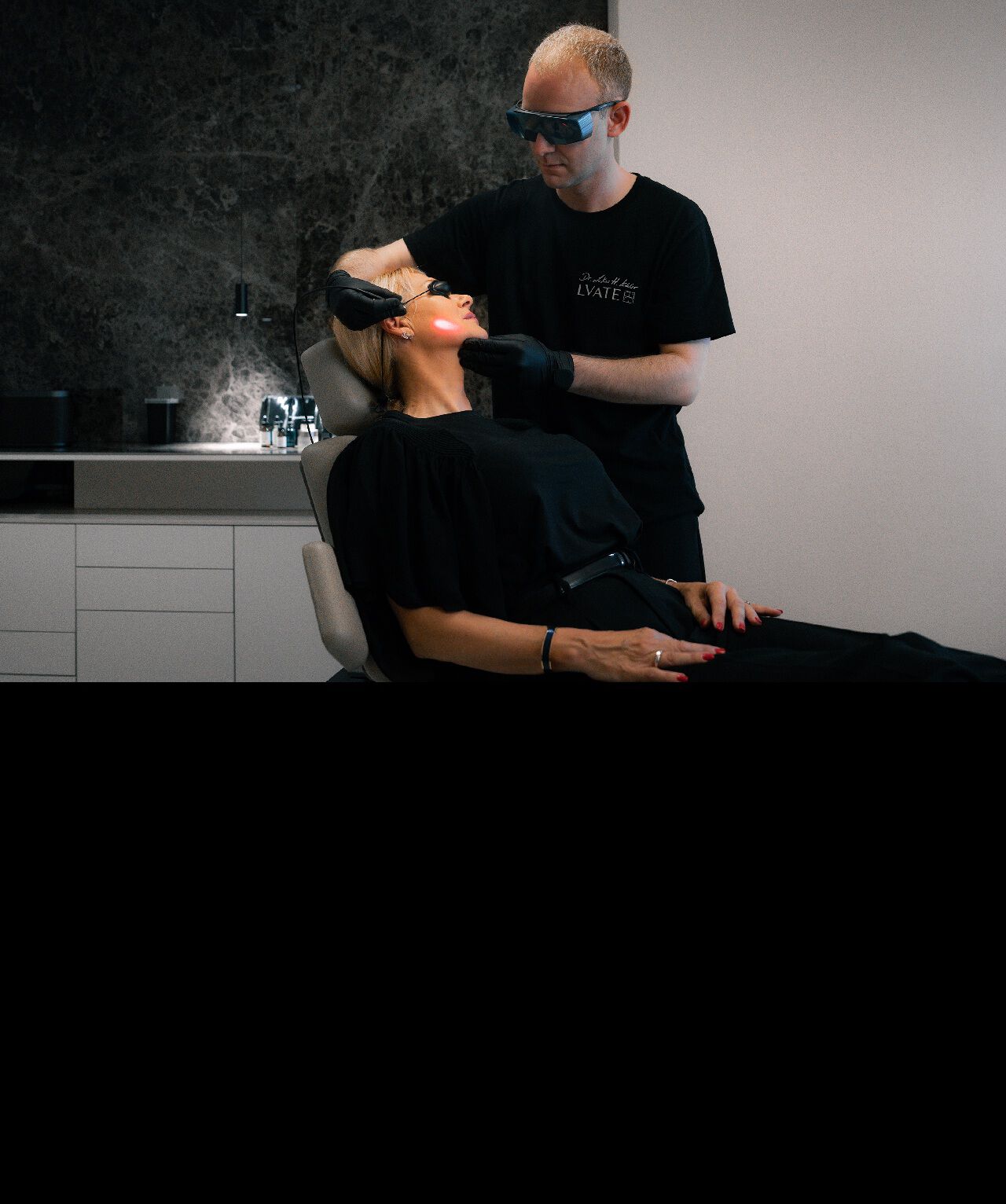 Un medecin en tenue de protection et lunettes de securite realise un soin au laser sur le visage d'une patiente allongee.​