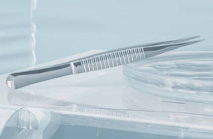 A close-up of a dental tool, specifically a tweezer, resting on a transparent surface, with soft lighting in a light blue environment.