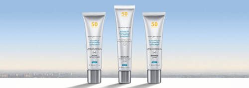 Three sleek tubes of skincare products are displayed against a soft gradient background that transitions from light blue to pale yellow, suggesting a serene skyline.