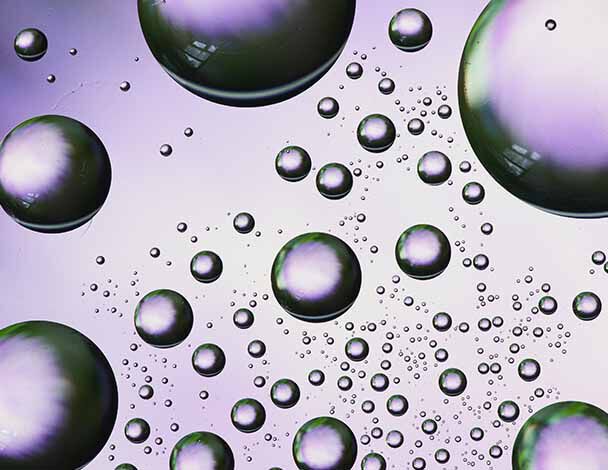 Close-up of iridescent water droplets floating on a textured surface, reflecting light in shades of green and purple.