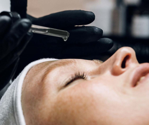 Nahaufnahme einer Frau mit ruhigem Gesicht, wahrend ein Kosmetiker in schwarzhandschuhen ein Serum auftragt.