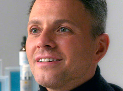 Un homme souriant avec des cheveux grisonnants, portant un pull noir, est assis dans un environnement lumineux.