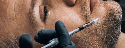 Un homme est allonge, recevant une injection sur le visage, avec des gants noirs visibles.
