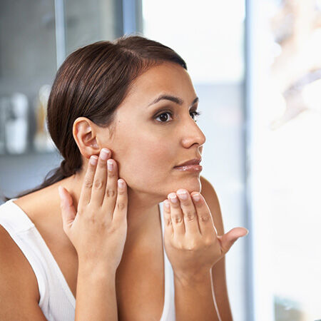 Une femme se regarde dans le miroir en touchant son visage, semblant preoccupee par sa peau.