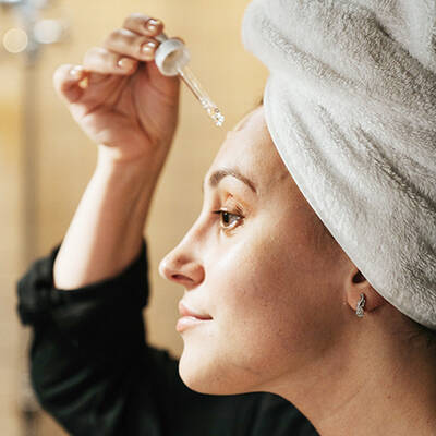 Une femme avec une serviette sur la tete applique un serum sur son visage, regardant dans un miroir.