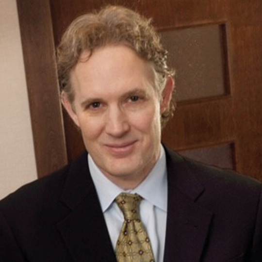 A smiling man with curly hair, wearing a suit and tie, stands in front of a wooden door.