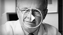 Ein älterer Mann mit Brille, der freundlich in die Kamera lächelt, vor einem unscharfen Hintergrund. Ein Videoknopf ist eingeblendet.